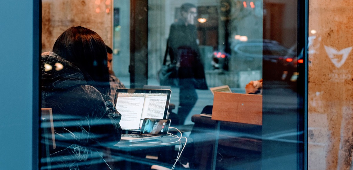 Symbolbild: Ein junge Frau durch ein Fenster fotografiert sitzt in einem Café und arbeitet an einem Notebook